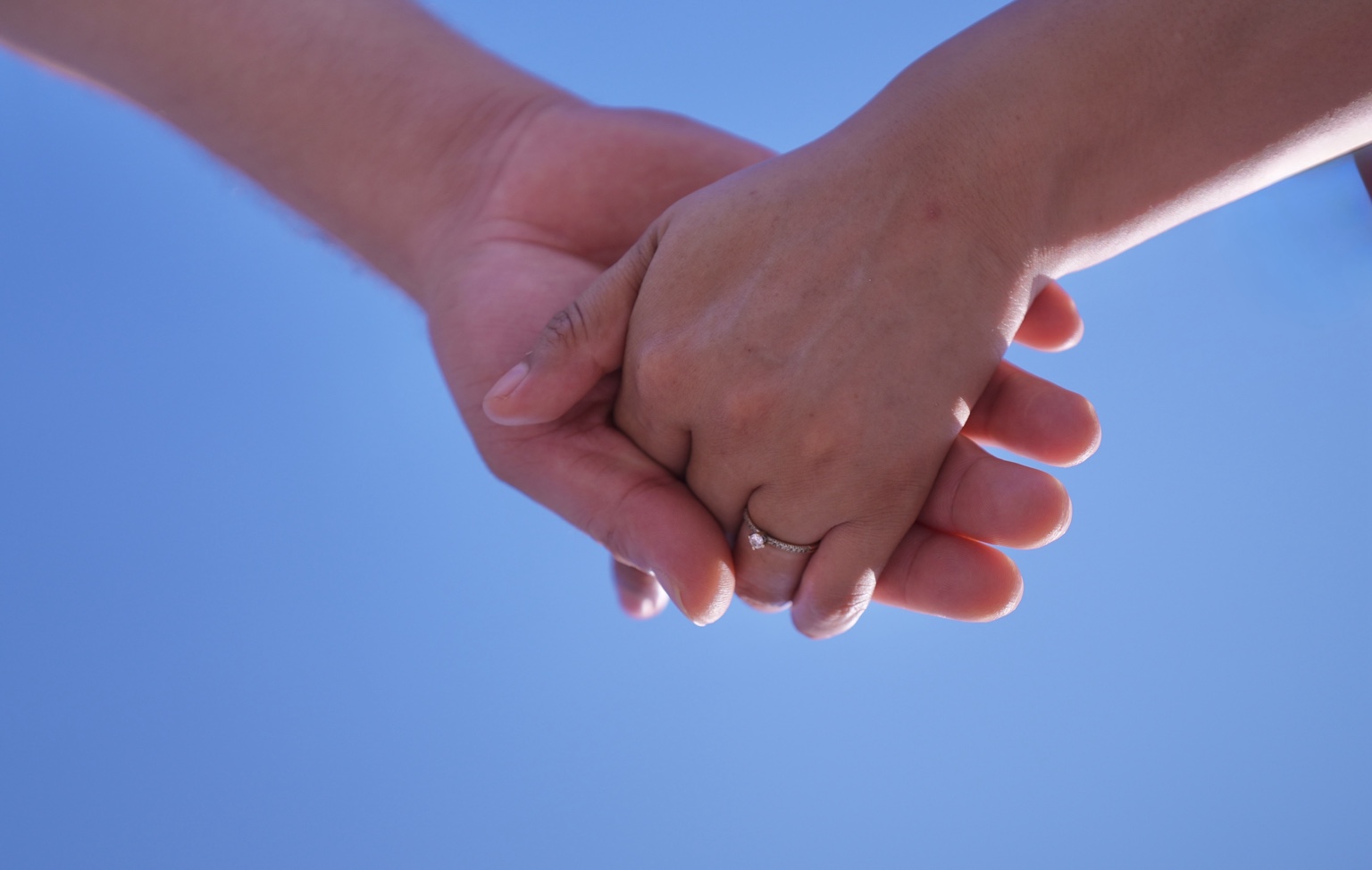 Primer plano de dos manos entrelazadas con un anillo de compromiso sobre un fondo de cielo azul